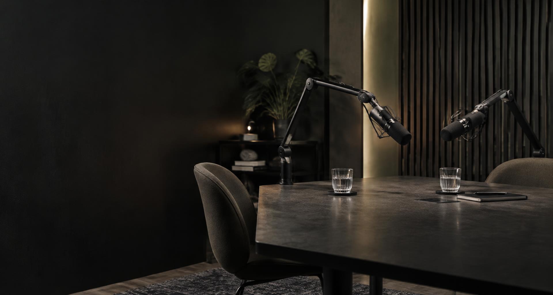 Leeres Podcast-Studio: zwei Mikrofone an Schwenkarmen über einem dunklen Tisch, zwei Stühle und warmes Licht an einer Holzlamellenwand